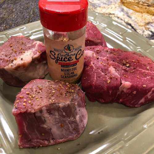 Four pieces of raw meat on a plate next to a jar of Manistee steak seasoning.