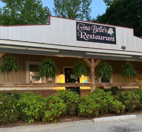 Exterior of Gina Belle's Restaurant, featuring a wooden facade and hanging plants, surrounded by greenery.