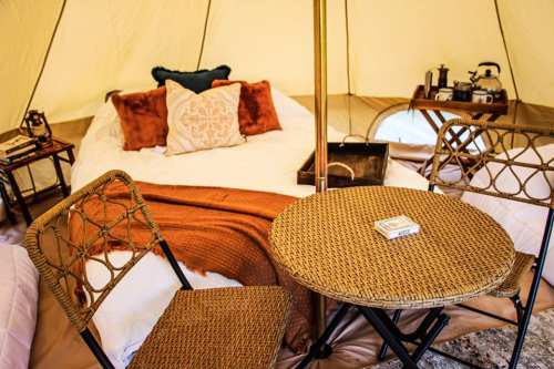 Cozy glamping tent interior with a bed, decorative pillows, a small table, and two chairs. Warm, inviting atmosphere.