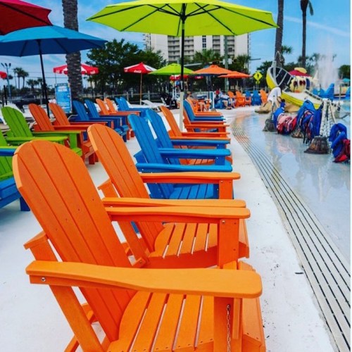 Colorful beach chairs in orange and blue line a poolside area, shaded by umbrellas under a clear blue sky.