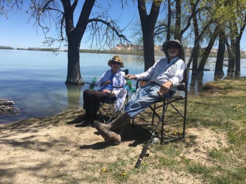 Two people sitting in chairs by a lake, surrounded by trees on a sunny day.