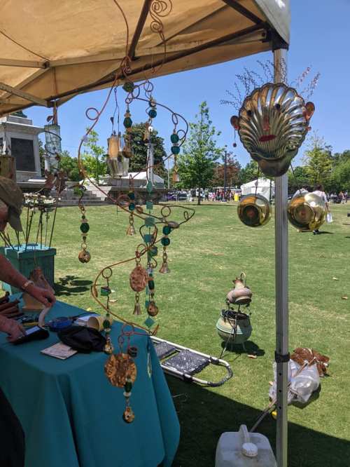 A craft booth featuring decorative metal sculptures and hanging ornaments, set up in a sunny outdoor park.