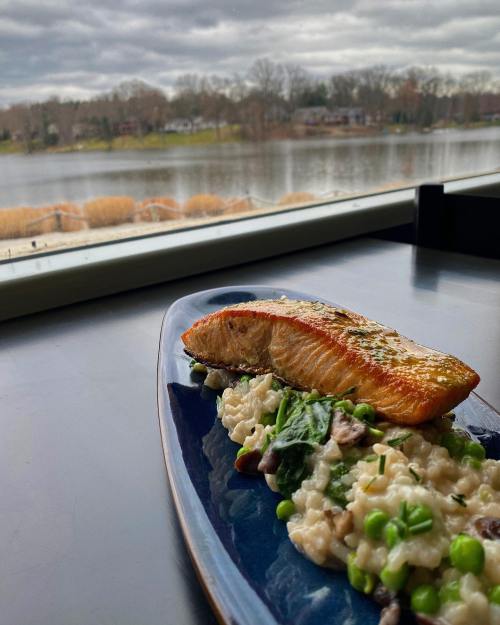 A plate of salmon on risotto with peas, set against a scenic lake view with cloudy skies.