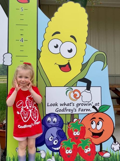 A young girl in a red shirt smiles next to a colorful farm sign featuring cartoon fruits and vegetables.
