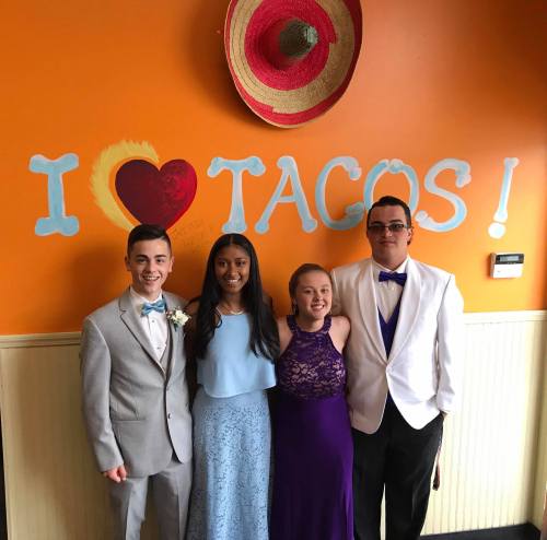 Four friends pose together in formal attire in front of a colorful "I ❤️ Tacos!" mural.