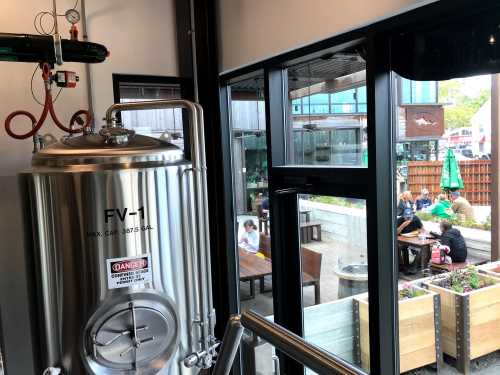 A brewing tank inside a brewery, with a view of outdoor seating and patrons enjoying their time.