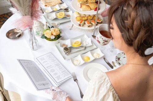 A woman in a vintage dress reads a menu at an elegant tea table set with pastries, tea, and floral decor.