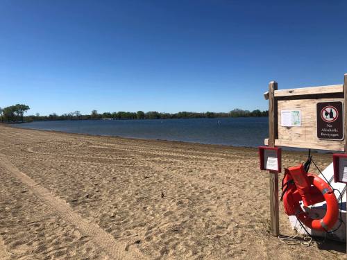 A sandy beach with a calm lake, featuring a lifebuoy and a sign warning against swimming. Clear blue sky above.