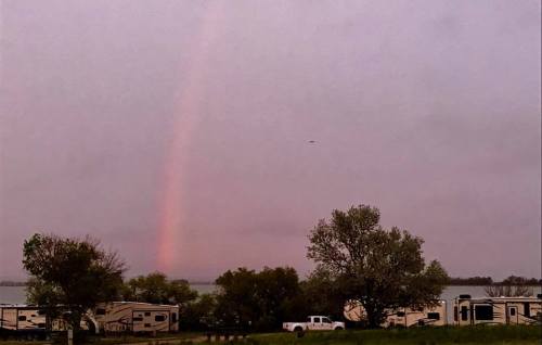 A rainbow arcs over a lake, with RVs and trees in the foreground under a cloudy sky.
