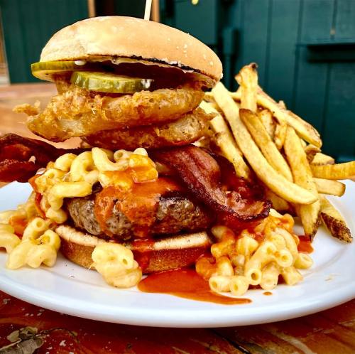 A towering burger with bacon, macaroni, and sauce, topped with pickles and onion rings, served with fries.