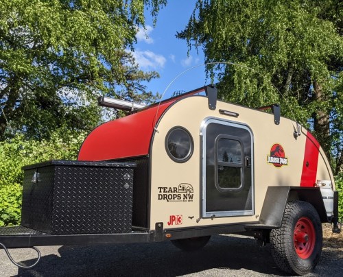 A small, retro-style teardrop trailer with a red and beige exterior, parked under trees.