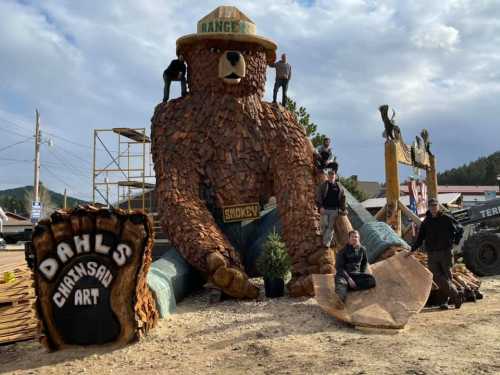 A large wooden bear sculpture with people working on it, surrounded by logs and tools, in a scenic outdoor setting.