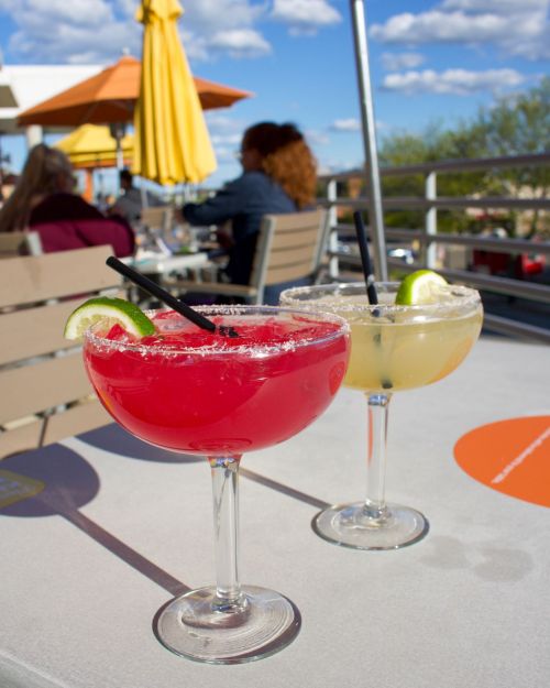 Two colorful cocktails in large glasses, one red and one yellow, with lime slices and salt rims, on a sunny outdoor table.