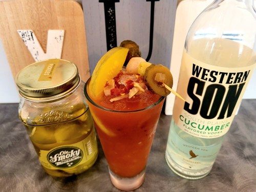 A glass of cocktail garnished with pickles and olives, alongside a jar of pickles and a bottle of cucumber-flavored vodka.