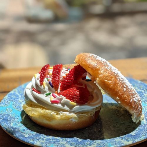 A colorful dessert featuring a donut topped with whipped cream, strawberries, and sprinkles on a decorative plate.