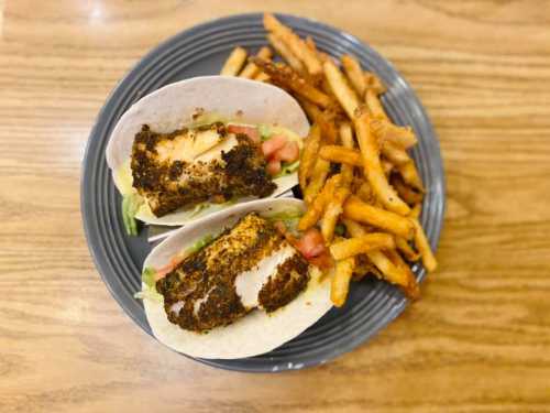 Two fish tacos with lettuce and tomato, served with a side of crispy fries on a gray plate.