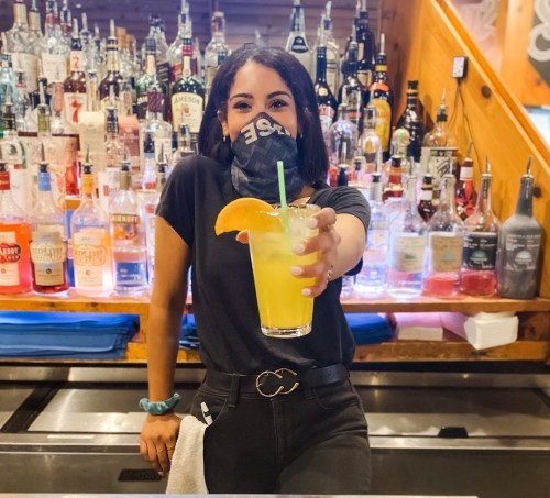 A bartender wearing a mask smiles while holding a colorful cocktail in front of a well-stocked bar.