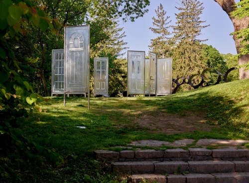 A serene outdoor scene featuring several transparent doors arranged on a grassy area, surrounded by trees.