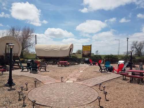 Outdoor seating area with colorful chairs, a stone path, and covered wagons under a blue sky with fluffy clouds.