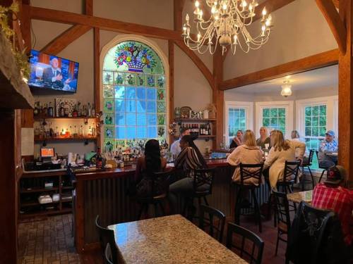 A cozy bar with a chandelier, stained glass window, and patrons enjoying drinks at the counter and tables.