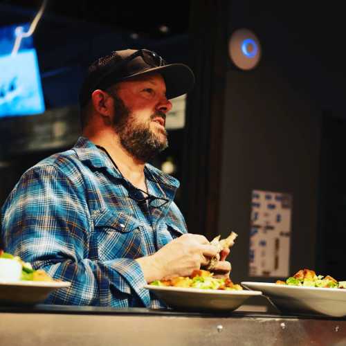 A man in a blue plaid shirt and cap sits at a table, holding a plate of food in a casual dining setting.