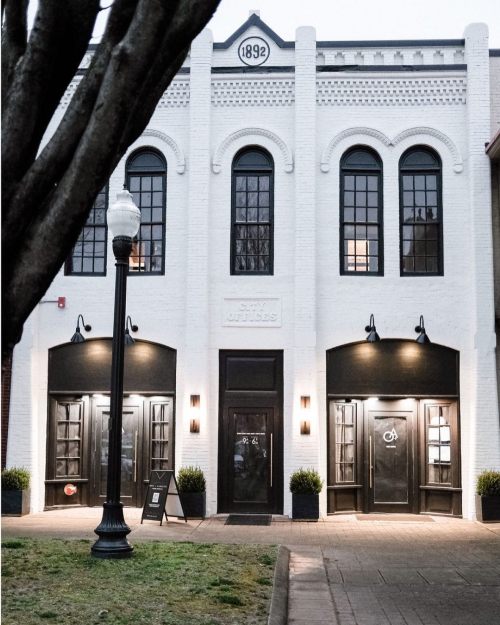 Historic white brick building with large black windows and doors, featuring a sign that reads "1892" above the entrance.