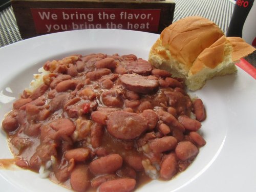 A plate of rice topped with red beans and sausage, accompanied by a soft bun, with a sign in the background.