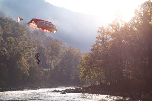 A parachutist descends over a river surrounded by trees and mountains, illuminated by sunlight.