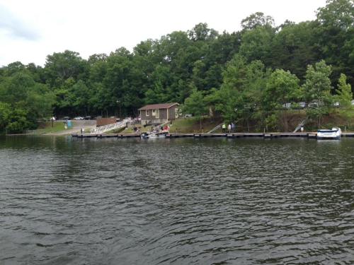 A serene lake scene with a dock, a small building, and people enjoying the outdoors among lush green trees.