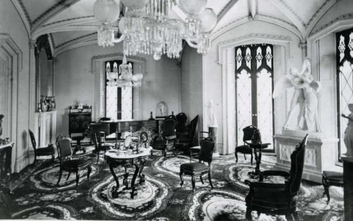 A vintage room with ornate furniture, a chandelier, and decorative sculptures, featuring a patterned carpet and large windows.