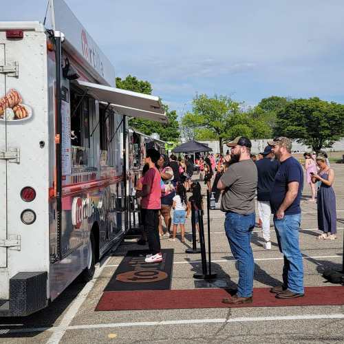 A food truck with people in line, surrounded by a crowd at an outdoor event on a sunny day.