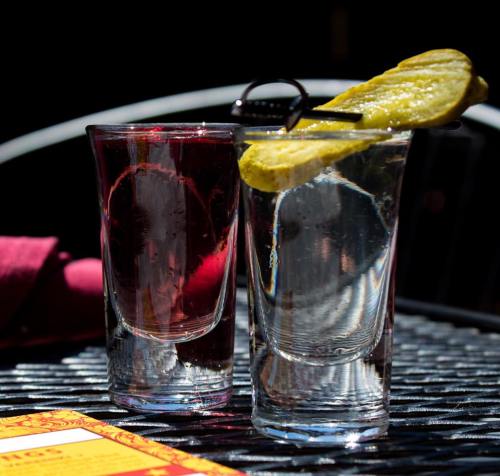 Two shot glasses on a table, one filled with a red drink and the other with clear liquid, each garnished with a pickle slice.