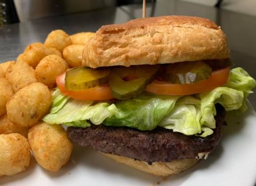A juicy hamburger with lettuce, tomato, and pickles, served with a side of golden tater tots on a white plate.