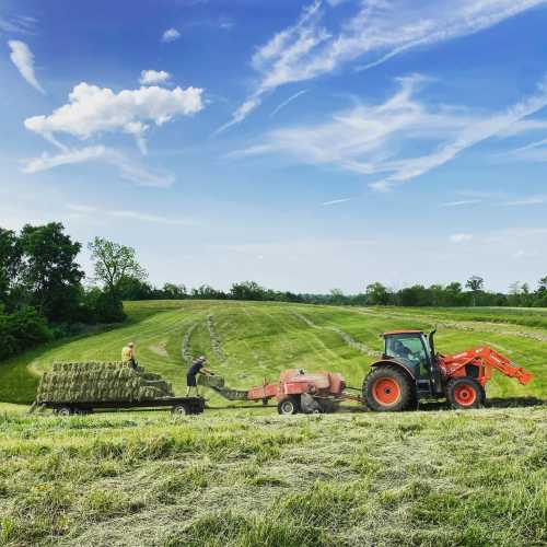 A tractor pulls a hay baler in a green field, with a person loading bales under a blue sky with scattered clouds.