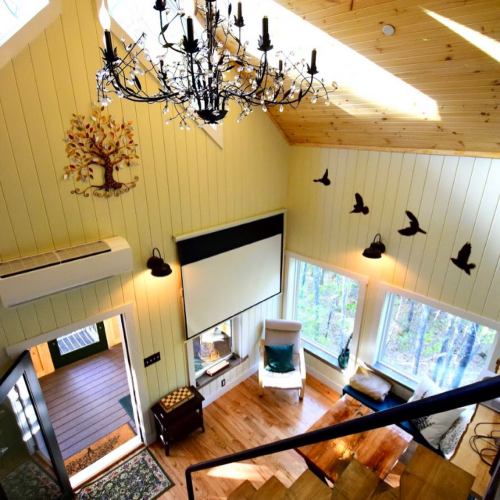 Cozy interior of a cabin with a chandelier, large windows, and wooden accents, featuring a tree wall art and birds.
