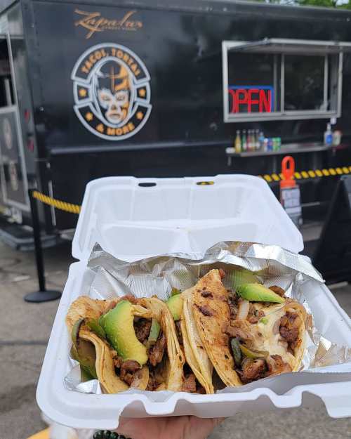 A hand holding a takeout container with three tacos, in front of a taco truck with an "Open" sign.