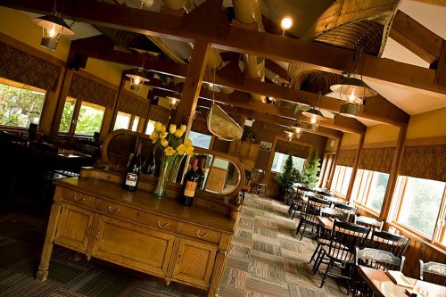 Interior of a cozy restaurant featuring wooden beams, tables, and a display of wine bottles with large windows.