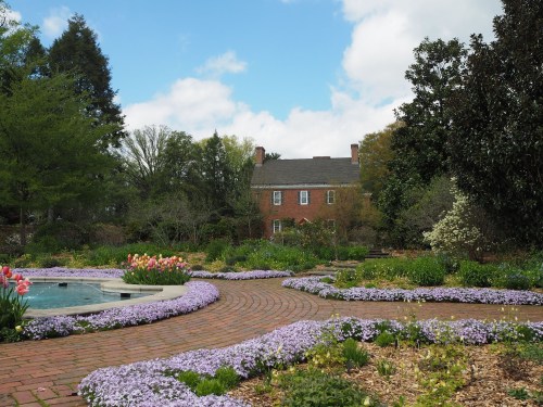 A scenic garden with blooming flowers, a winding brick path, and a house in the background under a partly cloudy sky.
