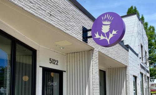 Exterior of a shop with a purple sign featuring a stylized thistle, located at 5122. Bright, sunny day.