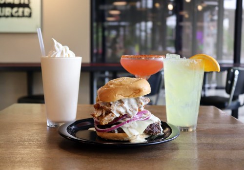 A plate with a burger topped with onions and sauce, accompanied by three drinks: a milkshake, a cocktail, and a margarita.
