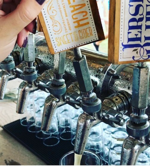 A hand holds a wooden sign above a row of beverage taps, with glasses ready to be filled underneath.