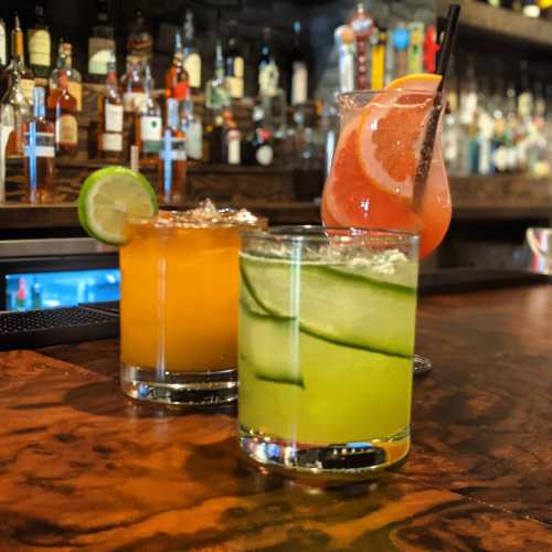 Three colorful cocktails on a bar: an orange drink, a green cucumber drink, and a pink grapefruit drink.