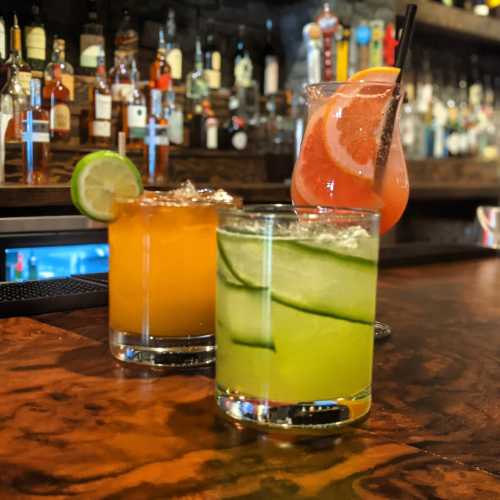 Three colorful cocktails on a bar: an orange drink, a green cucumber drink, and a pink grapefruit drink.