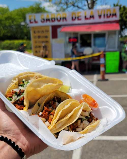 A hand holding a takeout container with three tacos, with a food truck in the background.