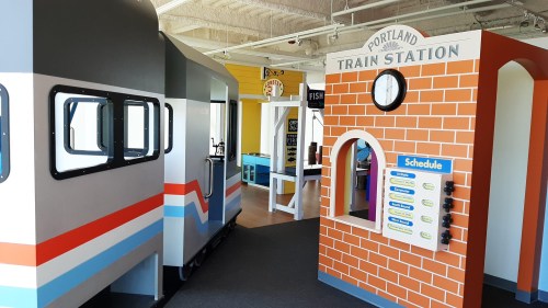 Colorful play area resembling a train station, featuring a train car and a brick wall with a schedule board.