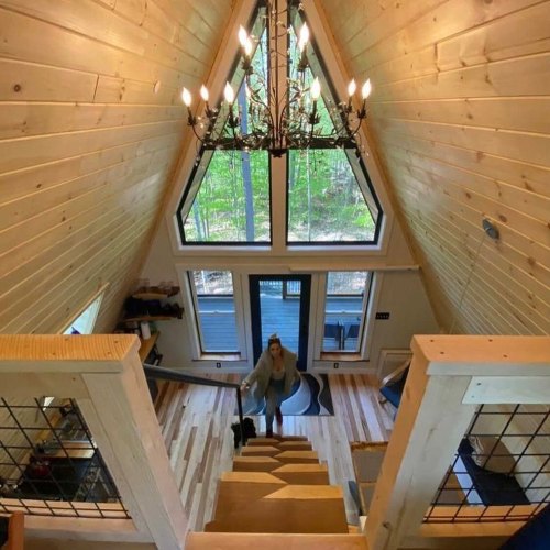 A cozy A-frame cabin interior with wooden stairs, large windows, and a chandelier, surrounded by greenery.