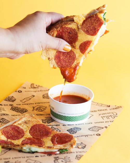 A hand holds a slice of pepperoni pizza dripping with cheese over a cup of marinara sauce on a yellow background.