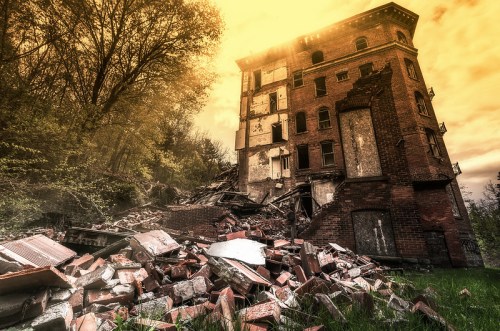 A dilapidated building with crumbling walls and debris, surrounded by trees under a golden sky.