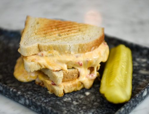 A grilled cheese sandwich with melted cheese oozing out, served with a pickle on a dark plate.