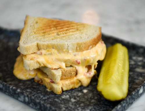 A grilled cheese sandwich with melted cheese oozing out, served with a pickle on a dark plate.